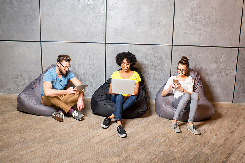 This image captures a casual setting with two women and one man, each comfortably seated on individual bean bags. They're positioned side by side in a relaxed atmosphere, likely a cozy workspace. Each of them is engrossed in their respective smartphones, perhaps working on personalization.