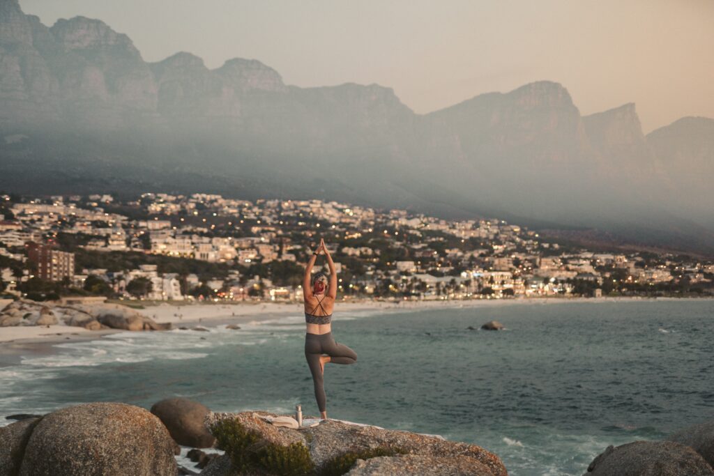 A woman getting exercise. She is balanced in Vrikshasana, or Tree Pose, on a rocky coastline overlooking the ocean. She is poised against a backdrop of serene, blue waters that stretch towards a coastal city skyline on the horizon. Her focused gaze and calm demeanor embody the tranquility of the surrounding landscape.