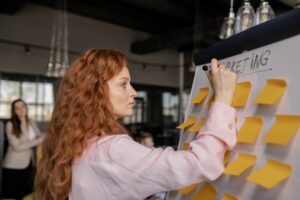 A woman in professional attire writing the word 'Marketing' in bold letters on a whiteboard, indicating a business strategy session or marketing planning specific to AIDA requirements.