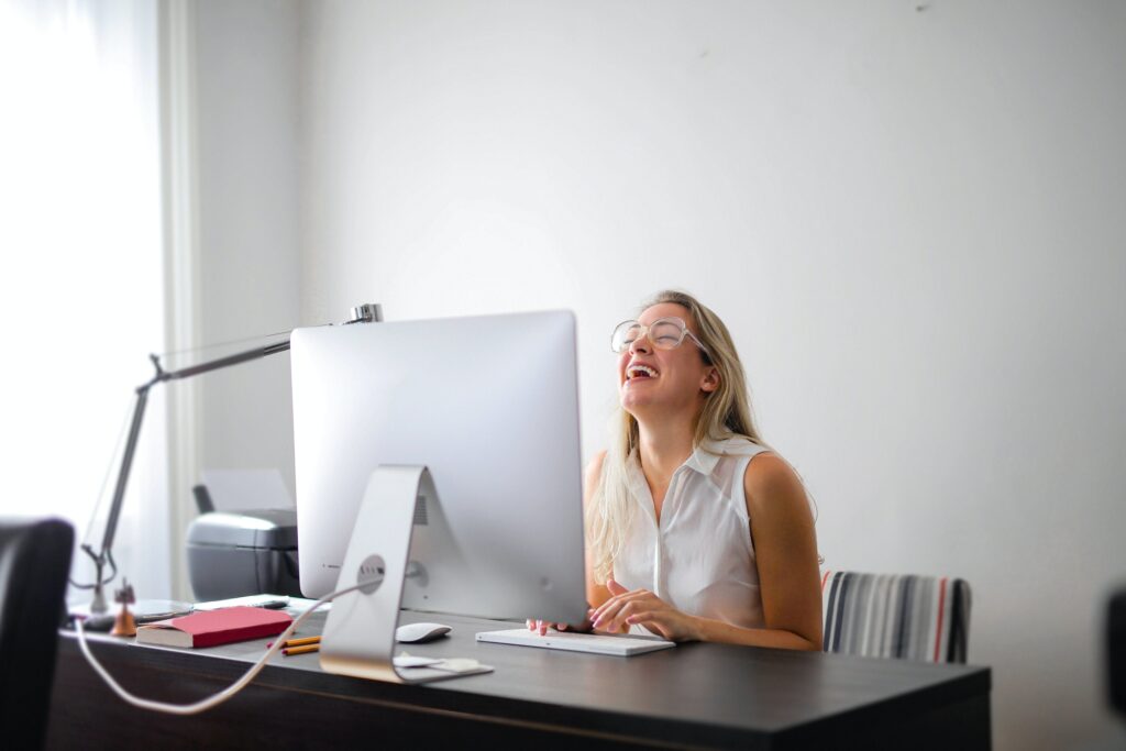 A joyful woman sitting at her computer in her office, typing and bursting into laughter at something on her screen. Her infectious laughter and engaged demeanor demonstrate the power and effectiveness of incorporating wit into the sales development process.