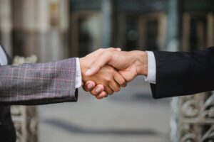 Two business professionals, one dressed in a black suit and the other in a plaid suit, extend their arms to engage in a firm handshake, signifying agreement, partnership, or a successful business sales development outcome.